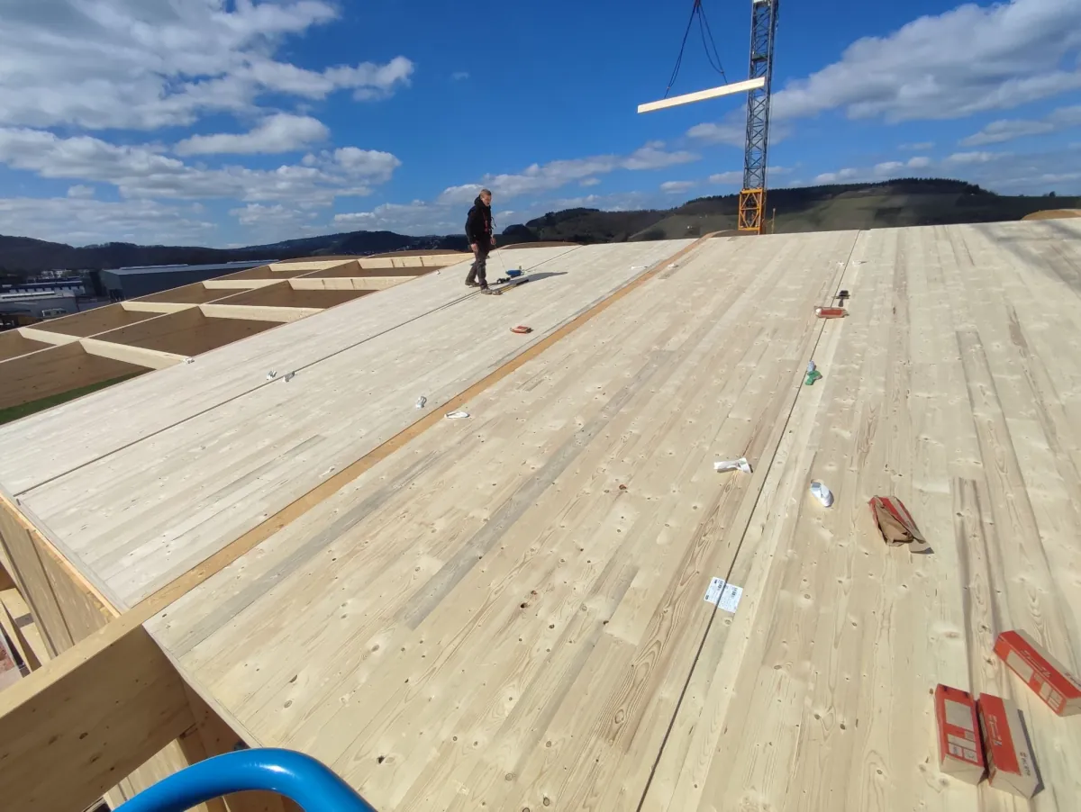 Zimmerer auf frisch verlegten Massivholz-Dachelementen, im Hintergrund hebt der Kran ein Holzbauteil ein